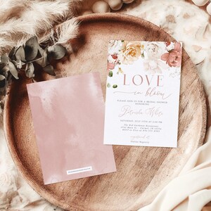 May include: A bridal shower invitation with floral accents and the words "Love in Bloom." The invitation is on a white background with a floral design in shades of pink, peach, and cream. A matching pink card is next to the invitation on a wooden tray.