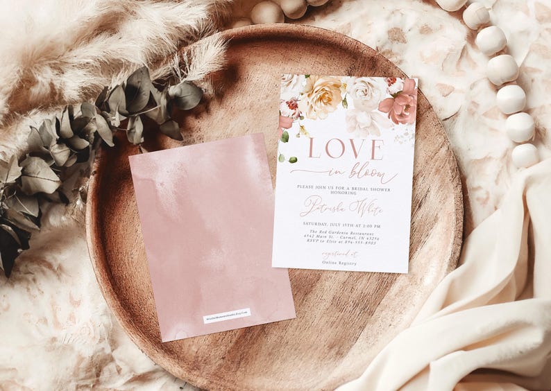 May include: A bridal shower invitation with floral accents and the words "Love in Bloom." The invitation is on a white background with a floral design in shades of pink, peach, and cream. A matching pink card is next to the invitation on a wooden tray.
