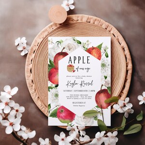 Może przedstawiać: Biała karta z czerwonym wzorem jabłka i tekstem "APPLE of our eye" i "Kaylin Russel" z kwiatową ramką. Karta jest zaproszeniem na baby shower z datą, godziną i miejscem wydarzenia.