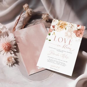 May include: A bridal shower invitation with floral accents and the words "LOVE in bloom." The invitation features a soft color palette of pink, white, and peach. Dried flowers and a pink card are also in the image.