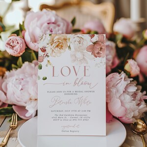 May include: A bridal shower invitation with the words "LOVE in bloom" in a delicate script. The invitation features floral accents in shades of pink, peach, and cream. The invitation is set on a white plate with gold cutlery and surrounded by pink peonies.