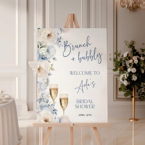 Peut inclure: Un panneau blanc sur un chevalet en bois avec le texte "Brunch + bubbly" et "Welcome to Ada's Bridal Shower April 12th". Le panneau est décoré d'éléments floraux bleus et blancs et de verres à champagne.
