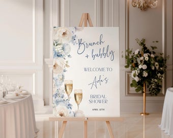 May include: A white sign on a wooden easel with the text "Brunch + bubbly" and "Welcome to Ada's Bridal Shower April 12th". The sign is decorated with blue and white floral accents and champagne glasses.