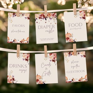 May include: Six cream-colored cards with fall floral and pumpkin designs are clipped to a white ribbon. Text on the cards includes "Favors," "Diaper Raffle," "Food," "Drinks," "Cards & Gifts," and "Guestbook." The cards are decorated with watercolor-style flowers.