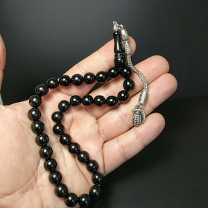 Può includere: Una collana di preghiera con perline nere e un ciondolo a forma di granata argentata. Le perline sono rotonde e lisce.
