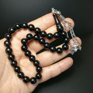 Può includere: Una collana di perline di preghiera nera con perline rotonde. Le perline sono tenute in una mano. La collana ha una catena d'argento e dettagli decorativi.
