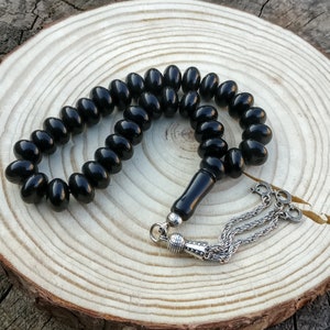 Può includere: Una collana di perline di preghiera nera con perline ovali e un nappa d'argento. Le perline sono infilate con un connettore nero e una catena d'argento. La collana è esposta su una superficie di legno.