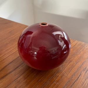 May include: A deep red, round ceramic vase with a smooth, glossy finish. The vase has a small opening at the top.