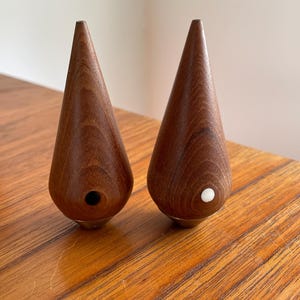 May include: A pair of wooden salt and pepper shakers in the shape of teardrops. The shakers are made of dark wood and have a white dot on the top of each shaker.