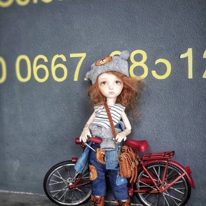 May include: A doll with red hair wearing a gray knitted hat with a bear face on it, a striped shirt, blue jeans, and brown boots. The doll is holding a brown leather bag and is standing next to a red bicycle.