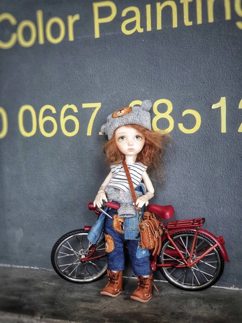 May include: A doll with red hair wearing a gray knitted hat with a bear face on it, a striped shirt, blue jeans, and brown boots. The doll is holding a brown leather bag and is standing next to a red bicycle.