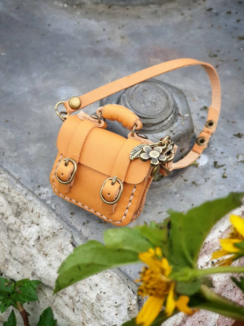 May include: A small, tan leather satchel with a strap and a metal clasp. The bag has two buckles on the front and a small metal charm attached to the strap.