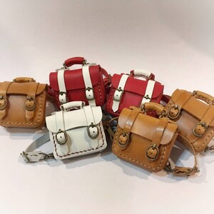 May include: Five miniature leather backpacks in different colors, including brown, white, and red. The backpacks have two buckles and a strap.