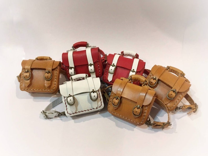May include: Five miniature leather backpacks in different colors, including brown, white, and red. The backpacks have two buckles and a strap.