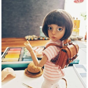 May include: A doll with brown hair and large blue eyes is wearing a pink and white striped shirt and a brown leather backpack. The doll is holding out its right arm and has a light brown hat on the table next to it.