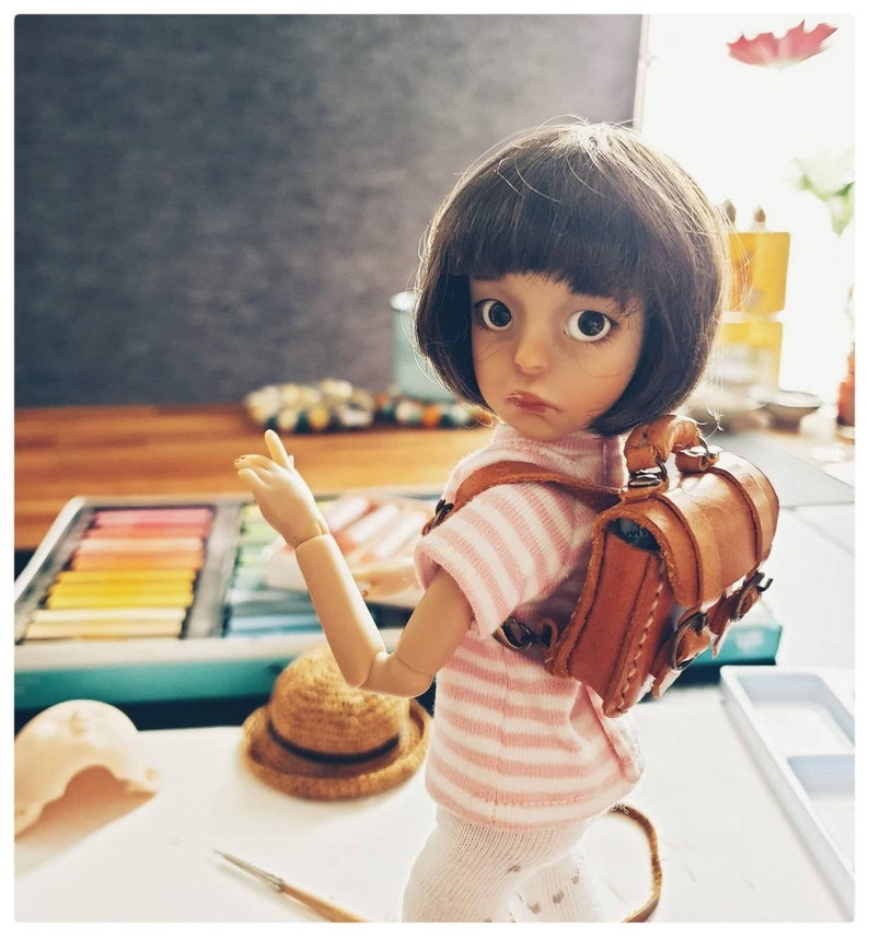 May include: A doll with brown hair and large blue eyes is wearing a pink and white striped shirt and a brown leather backpack. The doll is holding out its right arm and has a light brown hat on the table next to it.