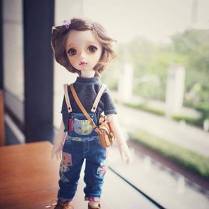 May include: A doll with brown hair wearing a grey shirt, blue overalls with floral patches, and brown shoes. The doll is holding a brown bag.