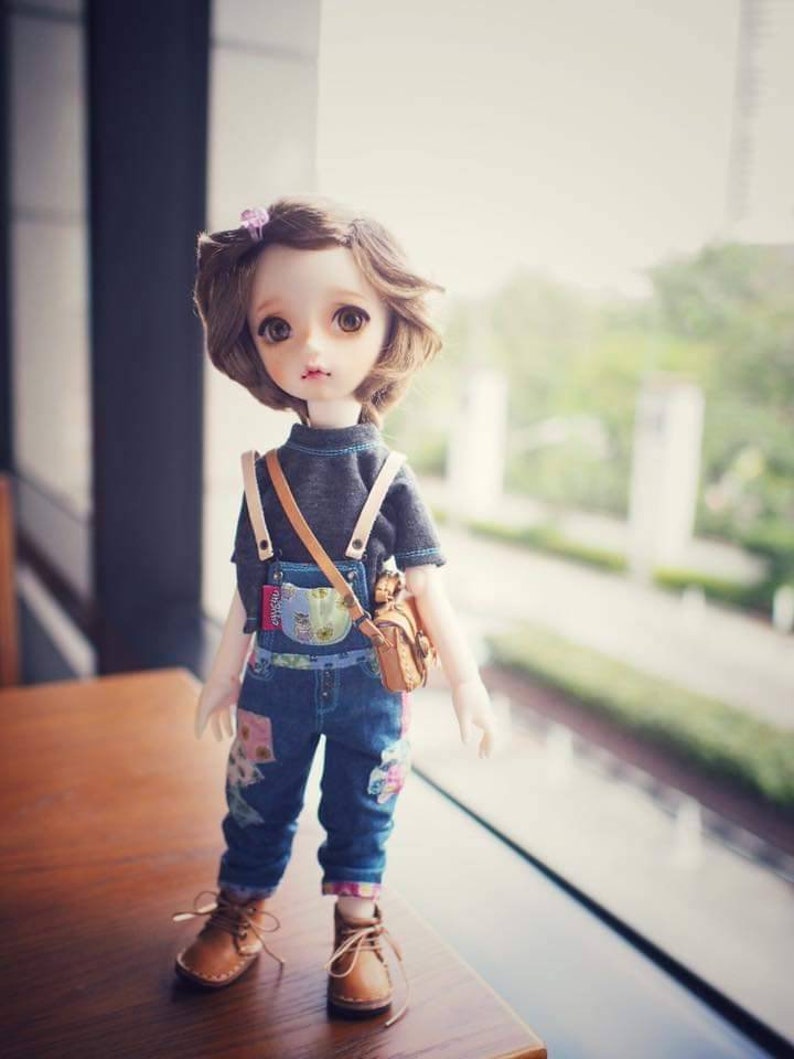 May include: A doll with brown hair wearing a grey shirt, blue overalls with floral patches, and brown shoes. The doll is holding a brown bag.
