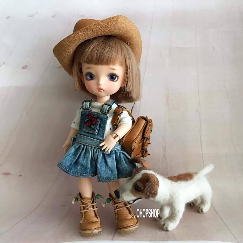 May include: A small doll wearing a brown cowboy hat, a white shirt with polka dots, a denim overall dress, and brown boots. The doll is holding a brown leather backpack and standing next to a small white and brown dog.