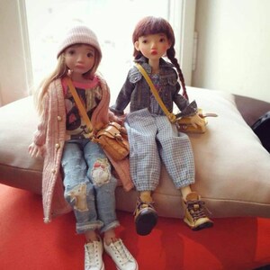 May include: Two dolls wearing casual outfits and carrying small bags. The doll on the left is wearing a pink cardigan, a white beanie, a graphic t-shirt, and blue jeans with patches. The doll on the right is wearing a denim jacket, a plaid dress, and brown boots.