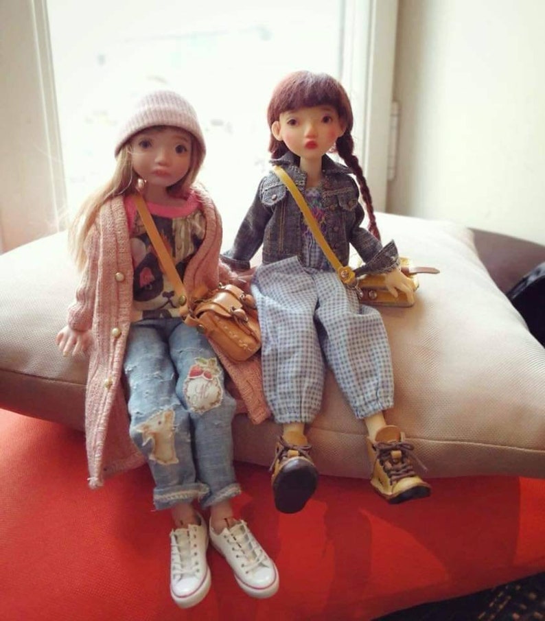 May include: Two dolls wearing casual outfits and carrying small bags. The doll on the left is wearing a pink cardigan, a white beanie, a graphic t-shirt, and blue jeans with patches. The doll on the right is wearing a denim jacket, a plaid dress, and brown boots.
