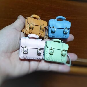May include: Four miniature leather satchel bags in different colors: brown, blue, pink, and green. Each bag has two buckles and a stitched design.