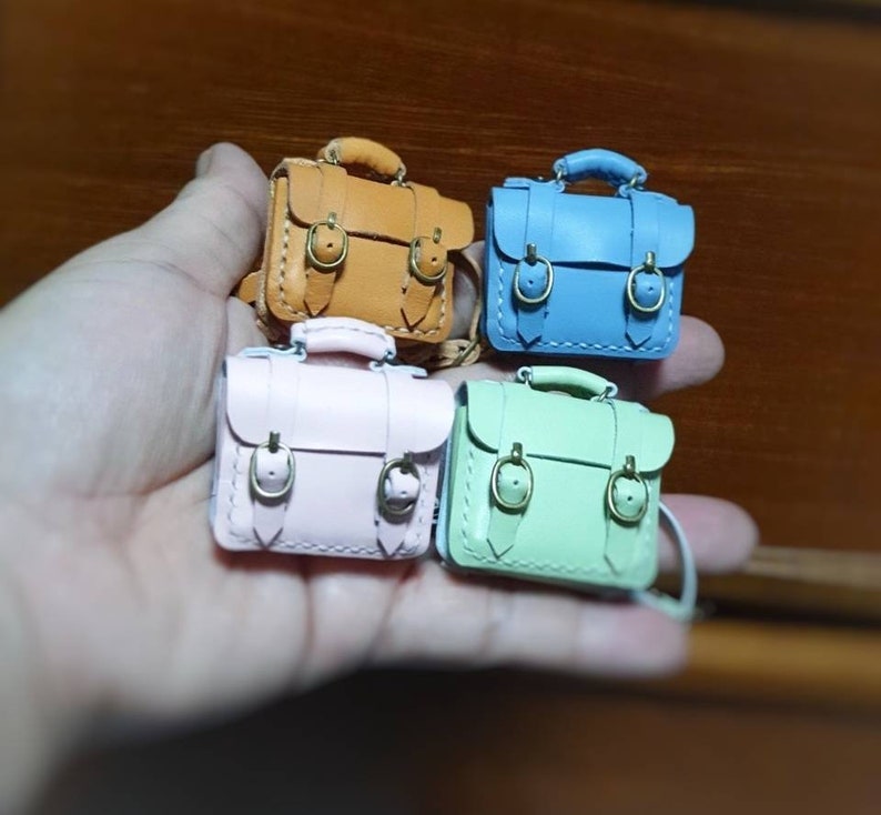 May include: Four miniature leather satchel bags in different colors: brown, blue, pink, and green. Each bag has two buckles and a stitched design.