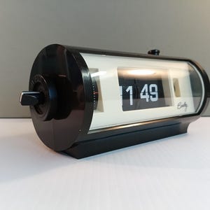 May include: A black and white flip clock displaying 1:49. The clock has a rectangular shape with rounded edges and a clear plastic cover. The clock has a black knob on the side and the brand name "Betty" is visible.