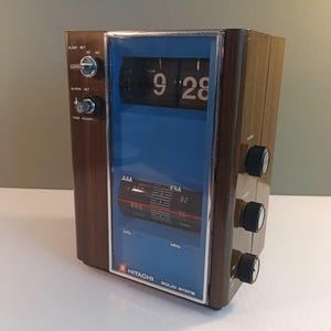 May include: A Hitachi solid state flip clock radio with a wood grain finish and a blue faceplate. The device features a digital flip display showing 9:28, an analog AM/FM radio tuner, a sleep set switch, an alarm set knob, and side volume controls.