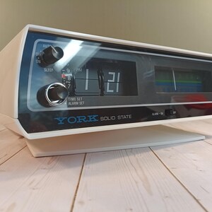 May include: A white York Solid State flip clock radio with a black front panel. The display shows the time 1:21 with a PM indicator. It features two black control knobs on the left, a radio frequency tuner on the right, and an alarm switch.