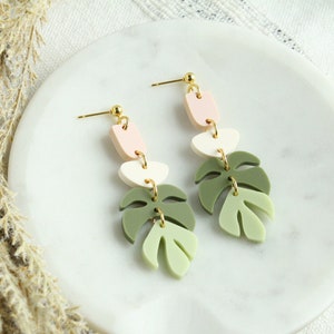 May include: A pair of dangle earrings featuring a layered design of pink, white, and green leaf shapes. The earrings have gold-toned accents.
