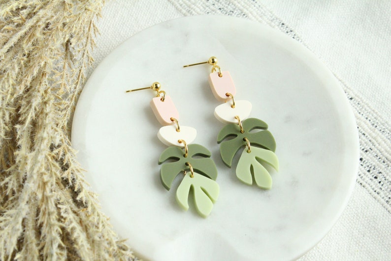 May include: A pair of dangle earrings featuring a layered design of pink, white, and green leaf shapes. The earrings have gold-toned accents.