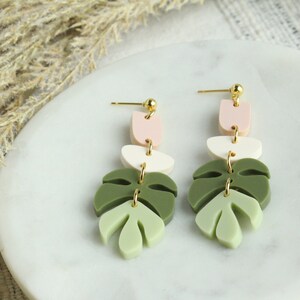 May include: A pair of dangle earrings featuring a layered design with pink, white, and green leaf shapes. The earrings have gold-toned accents.