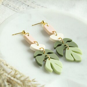 May include: A pair of dangle earrings with a pink, white, and green layered design. The earrings feature a monstera leaf shape.