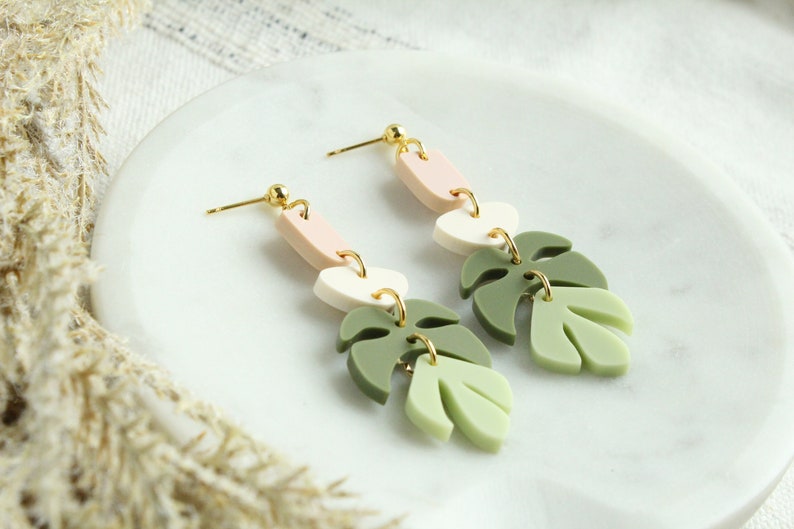 May include: A pair of dangle earrings with a pink, white, and green layered design. The earrings feature a monstera leaf shape.