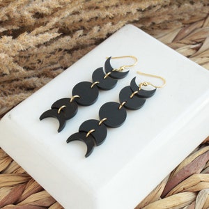 May include: A pair of black moon phase earrings with gold accents. Each earring features five crescent moon shapes connected by gold rings.