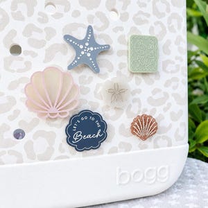 May include: A white bag with a beige leopard print pattern features several decorative charms: a blue starfish, a light green postage stamp with shell designs, a pink seashell, a sand dollar, a dark blue scalloped charm with the text Let's Go To The Beach, and a copper glitter seashell.