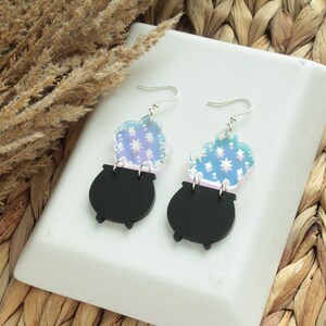 May include: A pair of black cauldron earrings with iridescent blue and white smoke accents. The earrings are made of acrylic and have silver hooks.