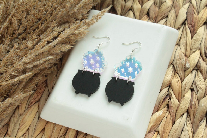 May include: A pair of black cauldron earrings with iridescent blue and white smoke accents. The earrings are made of acrylic and have silver hooks.
