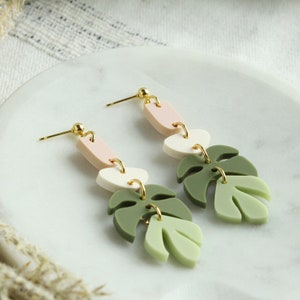 May include: A pair of dangle earrings with a green, white, and pink geometric design. The earrings feature a leaf shape and a gold hook.