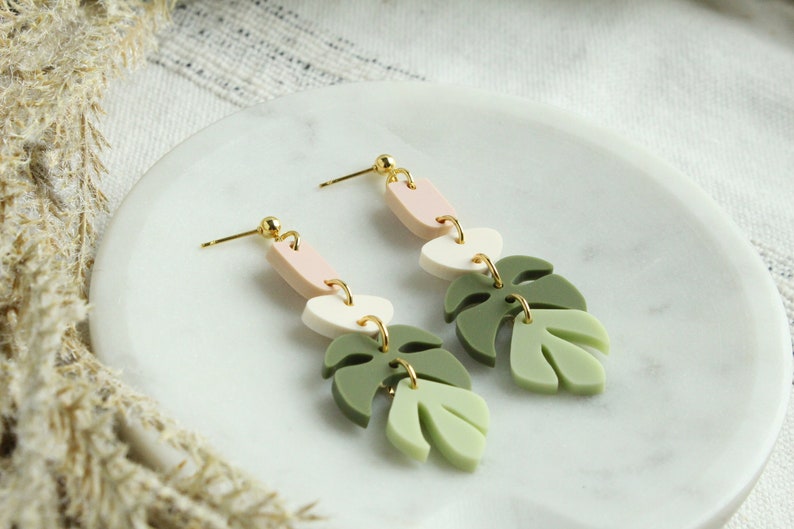 May include: A pair of dangle earrings with a green, white, and pink geometric design. The earrings feature a leaf shape and a gold hook.