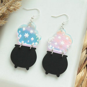 May include: A pair of iridescent earrings featuring a black cauldron with a swirling cloud of iridescent stars above it.