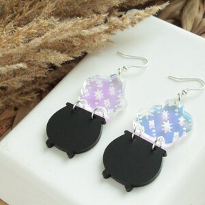 May include: A pair of iridescent black cauldron earrings with silver hooks. The cauldron is topped with a clear iridescent charm with a star pattern.