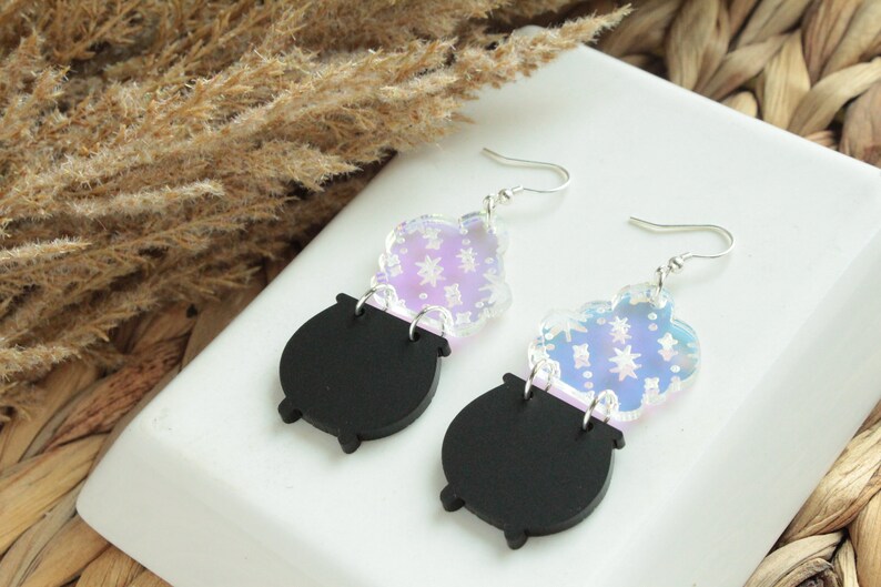 May include: A pair of iridescent black cauldron earrings with silver hooks. The cauldron is topped with a clear iridescent charm with a star pattern.