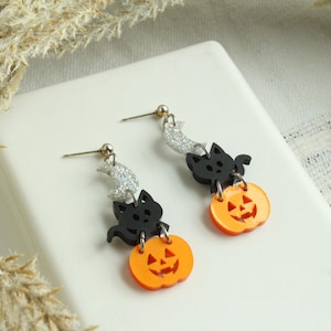 May include: A pair of Halloween-themed earrings featuring black cats and orange pumpkins. Each earring has a silver crescent moon charm, a black cat charm, and an orange pumpkin charm with a jack-o'-lantern face.