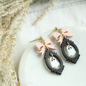 May include: A pair of Halloween-themed earrings. Each earring features a black frame with a white ghost design, a light pink bow, and gold-colored accents. The earrings are displayed on a white surface.