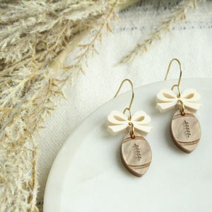 May include: A pair of earrings featuring a cream-colored bow and a football-shaped pendant. The pendant is light brown with etched details. The earrings have gold-colored hooks. The background includes dried plants and a white plate.