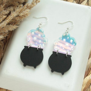 May include: A pair of iridescent earrings featuring a black cauldron with a swirling cloud of iridescent stars above it.