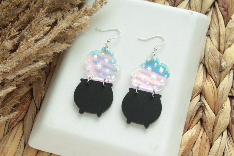 May include: A pair of iridescent earrings featuring a black cauldron with a swirling cloud of iridescent stars above it.