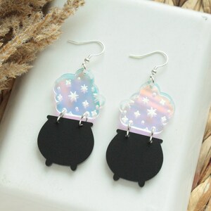 May include: A pair of iridescent earrings featuring a black cauldron with a swirling cloud of stars above it.